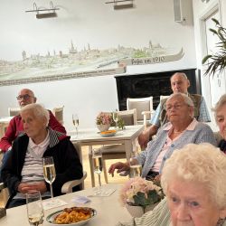 Eine Gruppe älterer Menschen sitzt in einem hellen Raum mit Snacks und Getränken, aufmerksam auf einen Vortrag.