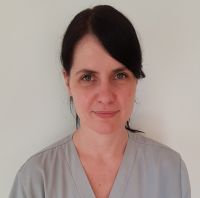 Eine Frau in grauer Arbeitskleidung blickt direkt in die Kamera vor einem neutralen Hintergrund.