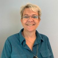 Eine lächelnde Person mit Brille trägt ein blaues Hemd vor einem neutralen Hintergrund.