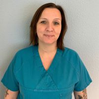 Eine Frau in blauer medizinischer Kleidung lächelt freundlich vor einer grauen Wand.