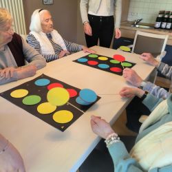 Senioren spielen gemeinsam ein Farbspiel mit bunten Kreisen auf einem Tisch in einem freundlichen Raum.