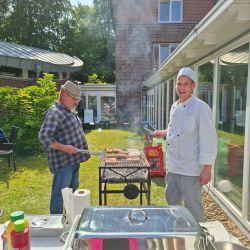 Ein Koch und ein Mann grillen Würstchen im Garten vor einem Backsteingebäude unter strahlend blauem Himmel.