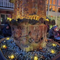 Ein beleuchteter Brunnen mit kunstvollen Reliefs ist von kleinen Kerzen umgeben, während Menschen im Hintergrund sitzen.