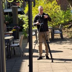 Ein Mann stellt eine Kamera auf einem Stativ im sonnigen Garten für eine Aufnahme ein. | © Name
