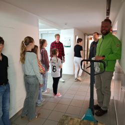 Eine Gruppe von Kindern und Erwachsenen wartet im Flur, während ein Mann mit einem Handhubwagen bereitsteht.