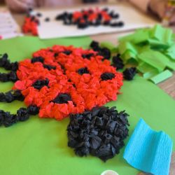 Eine kreative Bastelszene zeigt eine Marienkäfer-Dekoration aus buntem Papier auf grünem Hintergrund.