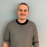 Ein Mann in einem gestreiften Pullover lächelt freundlich vor einer hellblauen Wand.