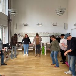 Eine Gruppe von Menschen steht im Kreis in einem Raum und macht gemeinsam eine Übung auf einem Holzboden.