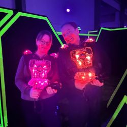 Zwei Personen in leuchtenden Westen spielen Laser-Tag in einer dunklen Arena mit neonfarbenen Barrieren.