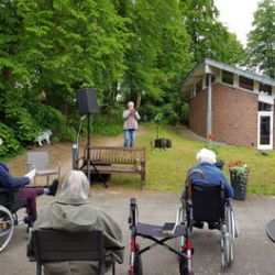 Im Freien hält eine Person eine Rede vor älteren Menschen in Rollstühlen und Stühlen, umgeben von grünen Bäumen.