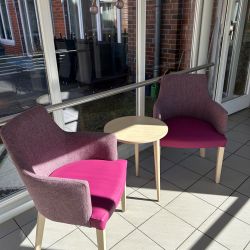 Zwei lila Sessel flankieren einen runden Tisch in einem sonnendurchfluteten Raum mit großen Fenstern.