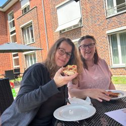 Zwei Frauen genießen bei Sonnenschein eine Mahlzeit an einem Tisch vor einem Backsteingebäude.