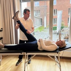 Ein Physiotherapeut dehnt das Bein einer Frau, die entspannt auf einer Behandlungsliege in einem hellen Raum liegt.