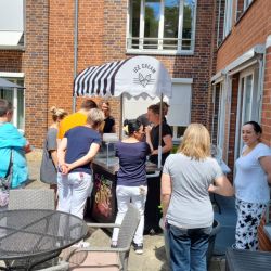 Menschen stehen an einem Eisstand vor einem Backsteingebäude und genießen das sonnige Wetter.