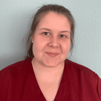 Eine lächelnde Frau in roter Arbeitskleidung steht vor einer hellblauen Wand und vermittelt Freundlichkeit und Professionalität.