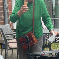 Eine ältere Frau im grünen Pullover genießt ein Eis im Freien, während ihr Rollator neben ihr steht.