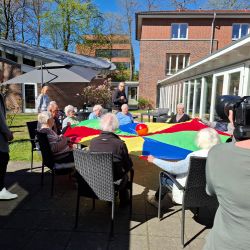 Senioren spielen mit einem bunten Fallschirm und einem Ball im Freien, umgeben von einem sonnigen Garten.