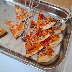 Ein Tablett mit übriggebliebenen Pizzastücken, belegt mit Gemüse und Käse, wartet auf hungrige Gäste. | © Name