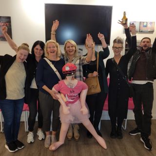 Eine fröhliche Gruppe posiert mit einer aufblasbaren Puppe vor pinken Stühlen und Kunstwerken an der Wand.