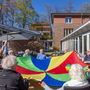Senioren spielen im Freien mit einem bunten Fallschirm und genießen den sonnigen Tag im Garten. | © Name