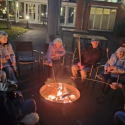Ältere Menschen sitzen abends um ein Lagerfeuer und grillen Würstchen auf langen Stöcken im Freien.