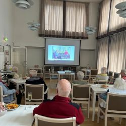 Ältere Menschen sitzen in einem Raum und schauen eine Präsentation auf einem großen Bildschirm an.