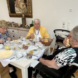 Drei ältere Frauen genießen ein gemeinsames Frühstück an einem liebevoll gedeckten Tisch in einem gemütlichen Raum.