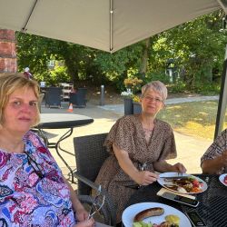 Drei Frauen genießen im Freien ein gemeinsames Essen mit Salat und Wurst unter einem schattenspendenden Sonnenschirm.