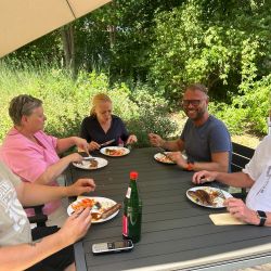 Fünf Personen genießen ein gemeinsames Essen im Freien unter einem Sonnenschirm bei sonnigem Wetter am Tisch.