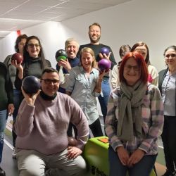 Eine Gruppe von Menschen posiert fröhlich mit Bowlingkugeln auf einer Kegelbahn für ein gemeinsames Foto.