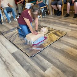 Eine Frau demonstriert die Herz-Lungen-Wiederbelebung an einer Übungspuppe vor einer Gruppe im Schulungsraum.