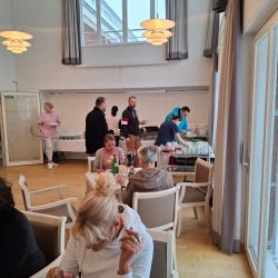 In einem hellen Speisesaal nehmen mehrere Personen an einem Buffet teil, während andere an Tischen essen.