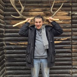 Ein Mann posiert fröhlich mit einem großen Geweih vor einer rustikalen Holzwand an einem Fotopunkt.