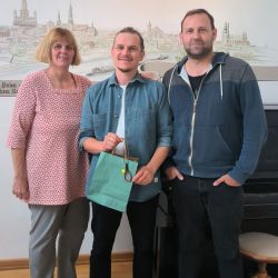 Drei Personen stehen lächelnd in einem Raum, während eine Person eine Geschenkverpackung hält.