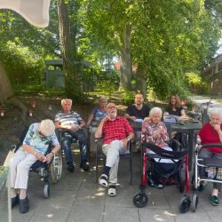 Ältere Menschen sitzen entspannt im Freien, umgeben von Bäumen, und genießen die sonnige Atmosphäre. | © Name
