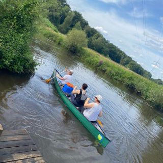 Drei Personen paddeln in einem grünen Kanu auf einem ruhigen Fluss, umgeben von üppiger Natur und klarem Himmel.