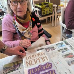 Eine ältere Frau mit Brille betrachtet ein Buch auf einem Tisch voller Zeitungen. | © Name