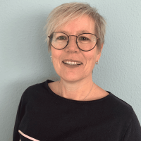 Eine lächelnde Frau mit kurzen, grauen Haaren und Brille trägt ein schwarzes Oberteil vor einer hellblauen Wand.