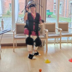 Eine Person balanciert im Raum mit Holzboden, trägt ein Exoskelett und steht zwischen Stühlen und bunten Markierungen.