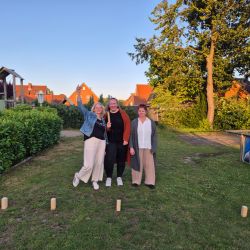 Drei Frauen stehen fröhlich im Freien, umgeben von Grün, während die Sonne untergeht. | © Name