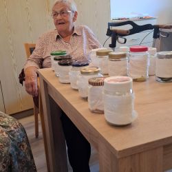 Eine ältere Frau sitzt an einem Tisch mit verschiedenen Gläsern, die sorgfältig aufgereiht sind.