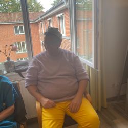 Eine Person in gelber Hose und lila Pullover sitzt entspannt vor einem Fenster mit Blick auf ein Backsteingebäude.