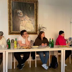 Vier Personen sitzen an einem Tisch und notieren sich während einer Besprechung etwas, umgeben von Getränken und Snacks.