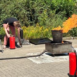 Eine Person bereitet mit einem Feuerlöscher einen Löscheinsatz vor, während Flammen aus einem Metallbehälter lodern.