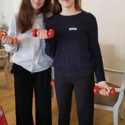 Zwei junge Frauen stehen mit Hanteln in einem gemütlichen Wohnzimmer und bereiten sich auf ein leichtes Fitness-Workout vor.