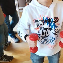 Ein Junge in einer weißen Kapuzenjacke hebt rote Hanteln, während er blaue Gewichte in einem belebten Raum an den Beinen trägt.