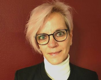 Eine Frau mit hellem Haar und Brille lächelt vor einem roten Hintergrund in die Kamera.