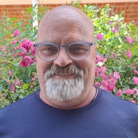 Ein lächelnder Mann mit Brille steht vor blühenden rosa Blumen im Hintergrund.