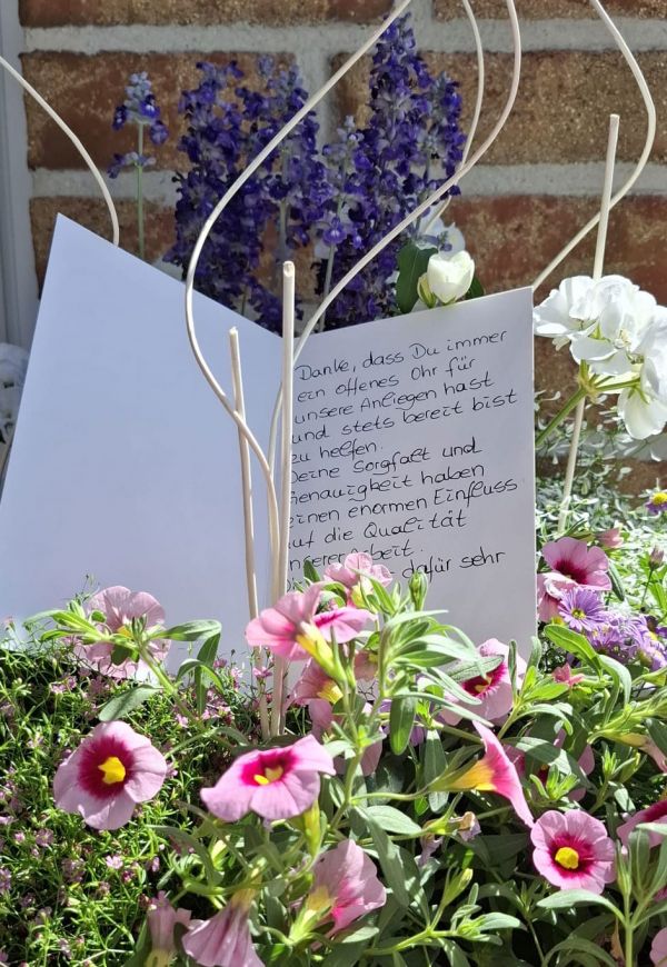 Ein handgeschriebener Brief steht dekorativ zwischen blühenden Blumen und Lavendel vor einer Ziegelmauer.