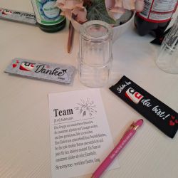 Ein Tisch mit einem Zettel über Teamarbeit, einem rosa Stift und Blumen, umgeben von Getränken und dekorativen Elementen.